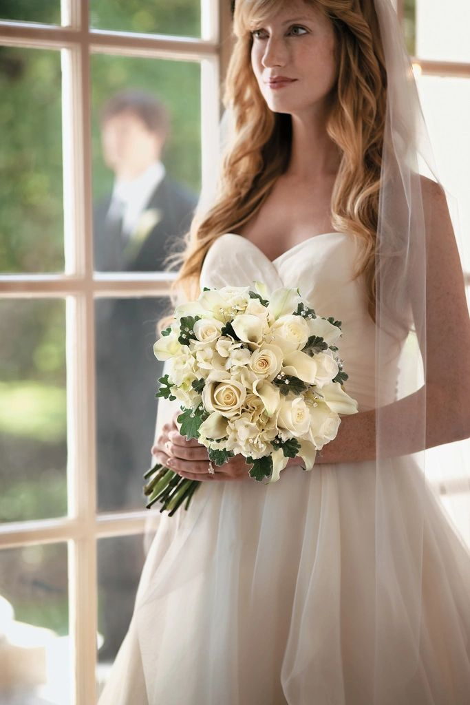 A bride holding a white bouquet, standing in window light before the ceremony.